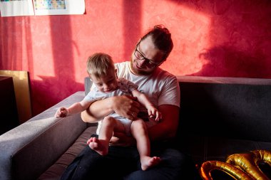 Farklılık abidesi babası, oğluyla kırmızı duvara yaslanmış gri koltukta eğleniyor. Bir adam küçük bir çocuğun ilk doğum gününü kutlar. Birinci balonların altın yazıtları. Ev hayatı partisi