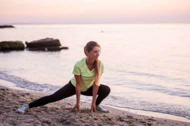 Spor giysili genç bir kadın sabahın erken saatlerinde gün doğumunda deniz kenarında mekik çeker. Uzun saçlı ince yapılı kız esniyor. Açık hava sporları, deniz kenarında. Spor dersleri, vücut bakımı, akıl sağlığı.
