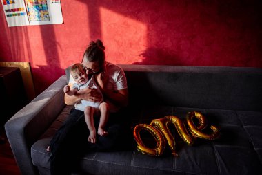 Farklılık abidesi babası, oğluyla kırmızı duvara yaslanmış gri koltukta eğleniyor. Bir adam küçük bir çocuğun ilk doğum gününü kutlar. Birinci balonların altın yazıtları. Ev hayatı partisi