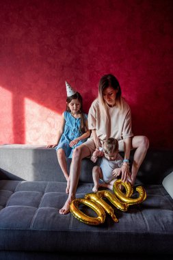 Yorgun genç anne, küçük çocuklarla kanepede oturuyor. Kırmızı duvar. Bekar bir aile, bebeklerin ilk yılını kutluyor. Folyo balonları bir gri koltukta. Evde yaşam tarzı partisi. Parti şapkalı küçük kız.