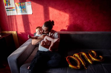 Farklılık abidesi babası, oğluyla kırmızı duvara yaslanmış gri koltukta eğleniyor. Bir adam küçük bir çocuğun ilk doğum gününü kutlar. Birinci balonların altın yazıtları. Ev hayatı partisi