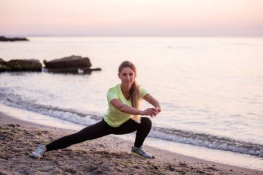 Spor giysili genç bir kadın sabahın erken saatlerinde gün doğumunda deniz kenarında mekik çeker. Uzun saçlı ince yapılı kız esniyor. Açık hava sporları, deniz kenarında. Spor dersleri, vücut bakımı, akıl sağlığı.