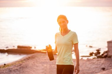 Antrenmanlar arasında mola veren spor giysili genç bir kadının portresi. Sporcu kadın denizde güneşin doğuşunu seyrediyor. Kız sabah rutininden sonra sette yürüyor. Açık hava egzersizi.