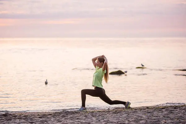Young woman in sportswear does sit-ups early in morning on seashore at sunrise. Girl of slim build with long hair is stretching. Outdoor sports by the water. Fitness classes, body care, mental health