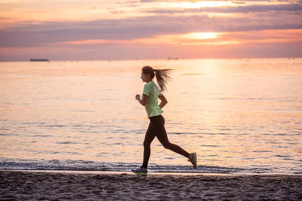 Young woman runner goes in for sports on the seashore at sunrise. Cardio load, fitness for fit, athletic girl. Beautiful sky. Outdoor exercise by water. Freedom of movement. Workout outside the home