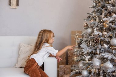 Şık görünümlü, hardallı, kadife pantolonlu, beyaz gömlekli küçük sarışın kız kırsal Noel ağacını süslüyor. Bebek kanepede oturuyor, arka planda makromlu gri duvar var. Moda bir iç mekan. Boşluğu kopyala