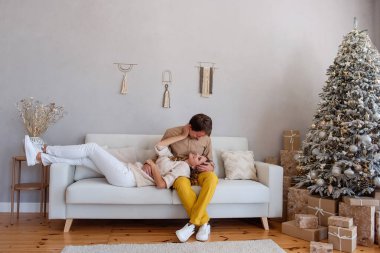 Sevgi dolu, neşeli bir çift Noel ağacının yanındaki koltukta hediyelerle beyaz bir kanepede oturuyor. Çılgın aşıklar tatillerini birlikte geçirirler. Şık, modaya uygun iç mekan, gri duvar, makro. Dokunulmazlık, samimiyet