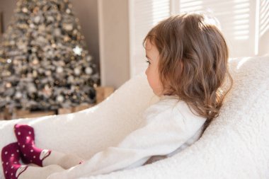 Üzgün küçük bir kız fotoğraflanmak istemez, kameraya arkasını döner, lensi eliyle kapatır. Zararlı, yaramaz çocuk beyaz yumuşak koltukta oturuyor Noel ağacının arka planında.