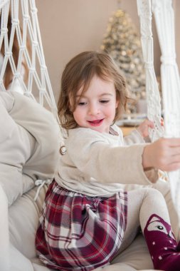 Evdeki makrome sandalyede salıncakta sallanan komik küçük kızın yakın plan portresi. Çocuk salıncak, oynaşma. Sıcak bir atmosfer. İç kısımda taşra tarzı. Çocuk kareli elbise giymiş.