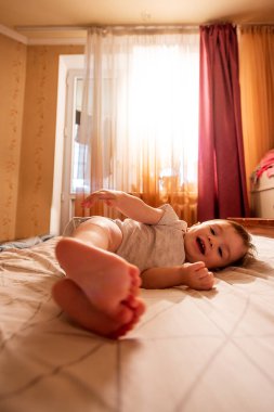 Bir yaşındaki bebek tulumu sırtüstü uzanıyor, sabahları güneş ışınlarıyla bacaklarıyla yatakta sallanıyor. Çocuk komik, gülümsüyor, keyfi yerinde. Bebeklere bakım ve hijyen. Çocuğun yakın plan portresi.