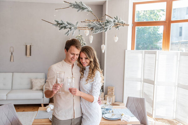 Close-up portrait of loving, young fashion couple with glasses of champagne in Christmas. Trendy interior in Scandinavian style with panoramic window and snow-covered branch. Lovers hugging, tactility