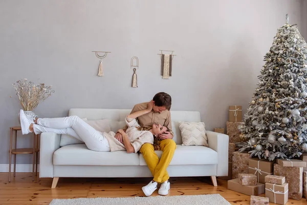 Loving, cheerful couple on a sofa near a Christmas tree with gifts sits on a white sofa. Crazy lovers spend the holidays together. Stylish, trendy interior, gray wall, macrome. Tactility, intimacy