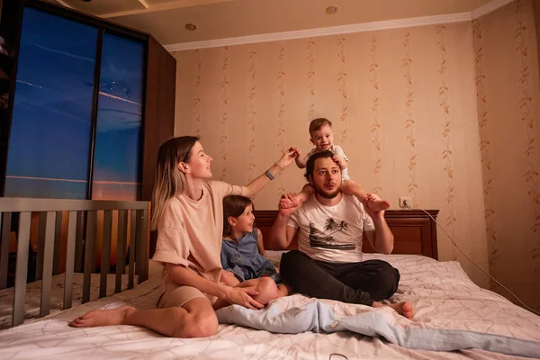 Funny portrait of diverse cheerful family on bed in morning. Stylish mother hugs daughter, father with glasses holds little son on shoulders. Parents children dance laugh fool around, have fun at home