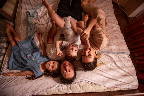 Top view of a cheerful family lying on the bed in the morning. Stylish mother and eldest daughter hug their father from both sides, the baby son crawls on the mans stomach. Domestic life, lifestyle