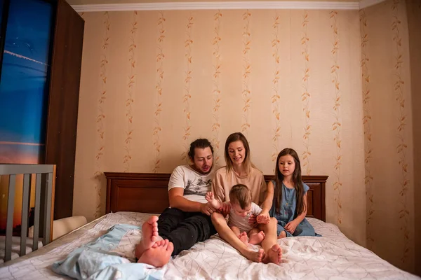 Long range shot of beautiful family on the bed in the morning in the sun from window. Tactile hugs of mother, father, daughter, son. Family weekend, relaxation at home. General view of family bedroom