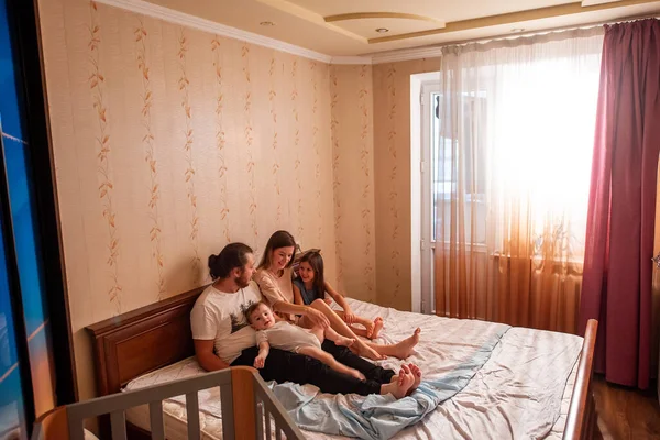 Long range shot of beautiful family on the bed in the morning in the sun from window. Tactile hugs of mother, father, daughter, son. Family weekend, relaxation at home. General view of family bedroom