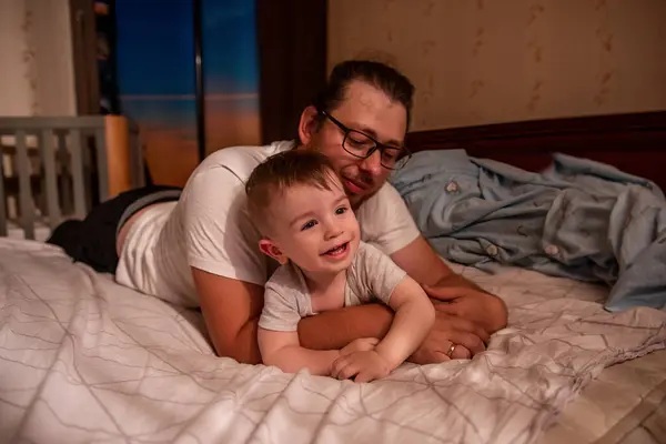 Diversity father with long hair and glasses hugs little son tenderly. Young man is fooling around with boy on bed in morning sun. Home games with parents. Family laughs happily. Lifestyle. Copy space