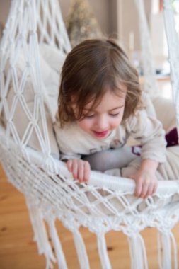 Evdeki makrome sandalyede salıncakta sallanan komik küçük kızın odaklanmış portresi. Çocuk salıncak, oynaşma. Sıcak bir atmosfer. İç mekanda kırsal biçim.