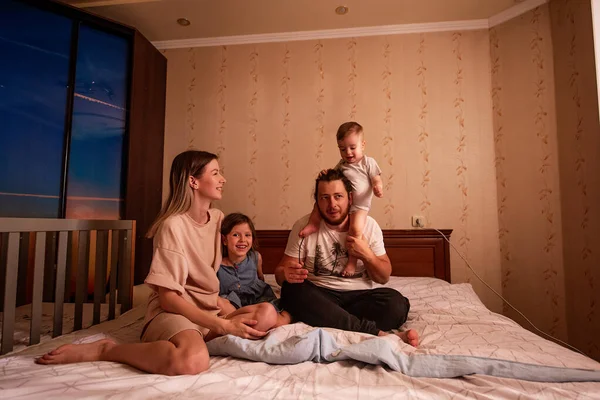 Funny portrait of diverse cheerful family on bed in morning. Stylish mother hugs daughter, father with glasses holds little son on shoulders. Parents children dance laugh fool around, have fun at home