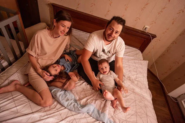 Top view of a cheerful family on the bed in the morning. Stylish mother, father with glasses tickle children, daughter and son in bodysuit laugh, fool around, have fun at home. Funny family lifestyle