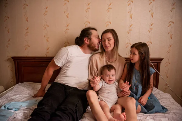 Funny, crazy portrait of a cheerful family on the bed in the morning. Tactile hugs of mother, father, daughter, son. Family weekend, relaxation at home. Young woman laughing while hugging children