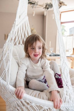 Evdeki Noel ağacının yanında hasır bir makrome salıncakta sallanan komik küçük bir kızın yakın plan portresi. Neşeli çocuk oyun oynuyor, dilini uzatıyor, oynaşıyor. Mutlu çocukluk