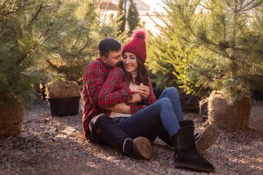 Sokak pazarında Noel ağacı fidelerinin arasında oturan kırmızı kareli gömlekli genç çiftin yakın plan portreleri. Erkek, örgü şapkalı kadını sevgiyle kucaklar. Sıcak kış tarihi.