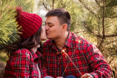 Kırmızı kareli gömlekli neşeli genç çift öpüşüyor, ormanda ellerinde neşeli maytaplar tutuyorlar. Erkek ve kadın Noel ağacı pazarında gülüyorlar. Mutlu aşıkların yakın plan portreleri. Yeni Yıl