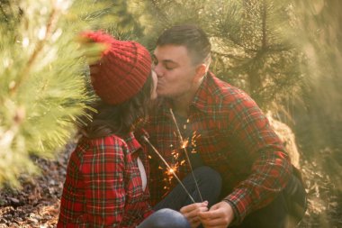 Çam ağaçlarının yeşil iğnelerinde, kırmızı kareli gömlekli mutlu çift ormanda neşeli maytaplar tutuyor. Şapkalı bir adam ve bir kadın Noel ağacı pazarında öpüşüyor. Sıcak Avrupa kışı. Boşluğu kopyala