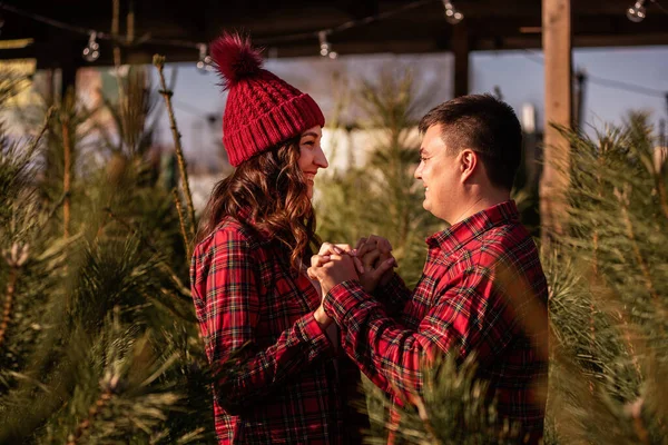 Cheerful couple in love in checkered red shirts, knitted hats hold hands among green Christmas tree market. Young man looks lovingly at beautiful woman without letting go of hands. Holiday atmosphere