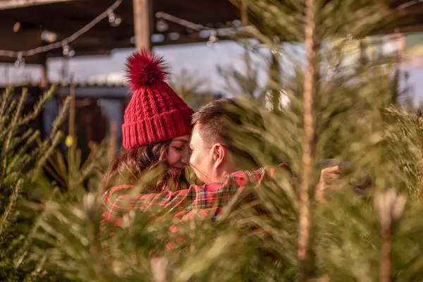 Beautiful couple in love in checkered red shirts, knitted hats hug tightly nose to nose face to face among green Christmas tree market. Young man laughs, kisses gorgeous woman. Holiday atmosphere