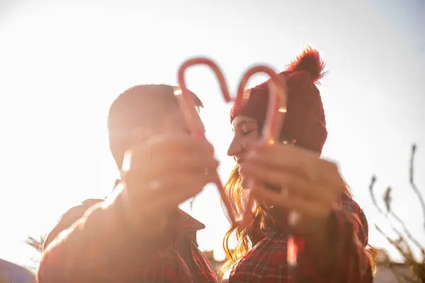 Couple in love in plaid shirts, knitted hats hold heart-shaped candy canes in hands near green market of Christmas trees. Man and woman kiss, laugh, have fun together. Tactility. Winter date
