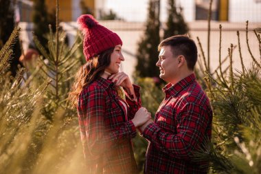 Kareli kırmızı gömlekli neşeli çift yeşil Noel ağacı pazarında el ele tutuşan örgü şapkalar. Genç adam ellerini bırakmadan güzel bir kadına sevgiyle bakar. Tatil atmosferi