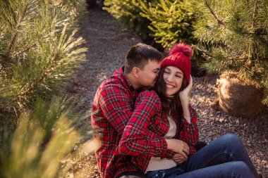 Ormanda gevşeyen, kırmızı kareli gömlekler giyen, Noel ağacı fidanlarının arasında oturan genç çiftlerin yakın plan portreleri. Erkek, örgü şapkalı kadını sevgiyle kucaklar, yanaktan öper.