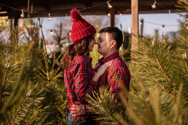 Funny couple in love in checkered red shirts knitted hats dances, spins among green Christmas tree market. Young man holds hands, kisses beautiful woman without letting go of hands. Holiday atmosphere