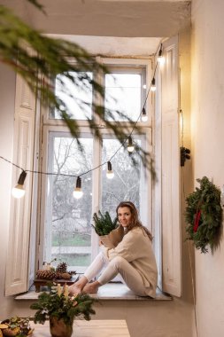 Dikey çekimde beyaz örgü süveter giyen, pencere kenarında oturan, çevre dostu kış buketi tutan, el işi kağıtta ladin dalları olan genç bir kadın. Otantik Noel dekorasyonu.