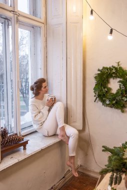 Genç şık bayan pencere kenarında oturur, bardaktan kakao içer, duvardaki DIY Noel çelengine bakar. Kış festivali atmosferi. Ev elbiseli bir kız çay ve kahve içer. Tatil lambaları