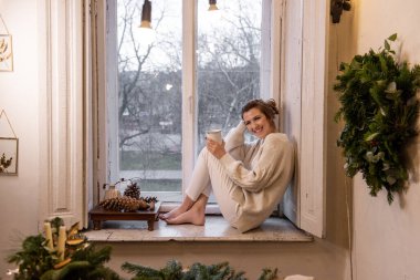 Genç, şık bir kadın pencerenin kenarında oturur, beyaz bardağı tutar ve güler. Duvarda el yapımı yeşil Noel çelengi asılı. Kış festivali atmosferi. Ev elbiseli kız çay, kahve, kakao içer.