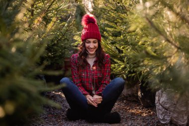 Elinde maytap olan, kırmızı kareli gömlek giyen, yeşil Noel ağacı pazarında örülmüş şapkası olan genç bir kadın. Kıvırcık saçlı kız yeni yılda güler ve sevinir. Doğa Tatili hissi. Boşluğu kopyala