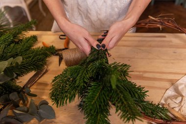 Kadın ellerinin doğal ladin, çam, okaliptüs dallarından Noel çelengi yapmasının üst görünümü. Kız yeşil dalları sicimle Kraft üssüne sarıyor. Kurutulmuş portakallar ahşap masada yatıyor.