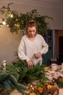 Genç bir kadın doğal ladin, çam ve okaliptüs dallarından Noel çelengi yapıyor. Kız yeşil dalları sicimle Kraft üssüne sarıyor. Kurutulmuş portakallar ahşap masada yatar. İskandinav iç mimarisi