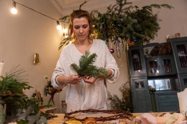 Genç kadın evde doğal malzemelerden el yapımı Noel çelengi yapıyor. İskandinav iç mimarisi olan otantik bir daire. Çiçekçiden talimat. Canlı çam iğneleriyle süslenmiş. Boşluğu kopyala