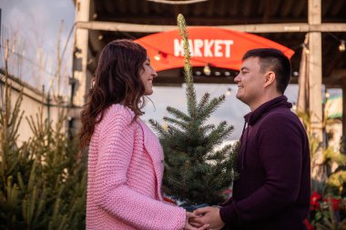 Birbirine aşık çiftler Noel ağacını açık hava pazarından seçer. Genç bir adam ve güzel bir kadın birbirlerine bakıyorlar, ellerinde canlı Noel ağacı olan bir küvet tutuyorlar. Tatil süslemeleri için çevre dostu seçim.