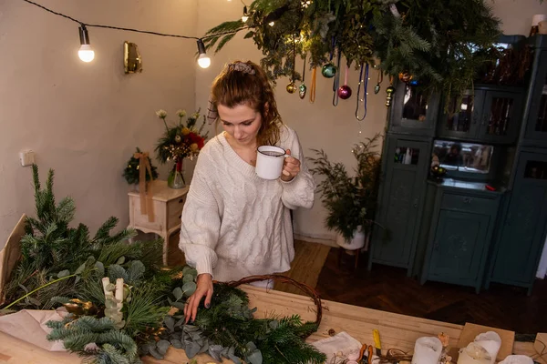 Young woman florist workshop makes Christmas wreath, drinks hot tea from white cup during break between work. Small business during holidays. Winter atmosphere. Blogger leads tutorial on social media