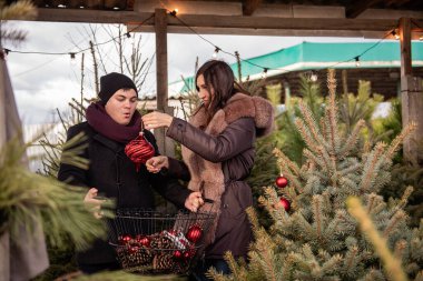 Komik çift, markette Noel ağacını kırmızı toplarla süslüyor. Genç adam ve kadın arka bahçedeki köknar ağacına oyuncaklar asarak rahat, şenlikli bir ortam yaratıyorlar. Adam sepeti tutuyor.