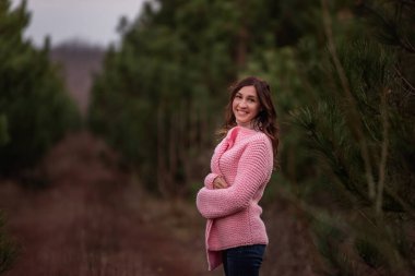 Pembe örgü süveter giyen genç ve güzel bir kadın, kozalaklı ormanların arasında eğleniyor. Kış doğada yürür. Doğal, botanik geçmişi. Kırsalda moda kıvırcık kız. Yaşam biçimi