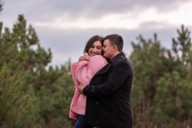 Yeşil kozalaklı ormanda sarılan mutlu çiftin yakın plan portresi. Genç adam kıvırcık saçlı pembe kazaklı kadının etrafına özenle siyah bir ceket sardı. Kış nişanı, sonbahar düğünü. Evlilik.