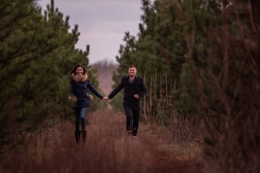 Ormanda el ele tutuşarak koşan mutlu çift. Siyah ceketli bir adam ve örgü kazaklı genç bir kadın çam parkında kovalamaca oynuyorlar. Kış doğası gereği. Sevgililer Günü