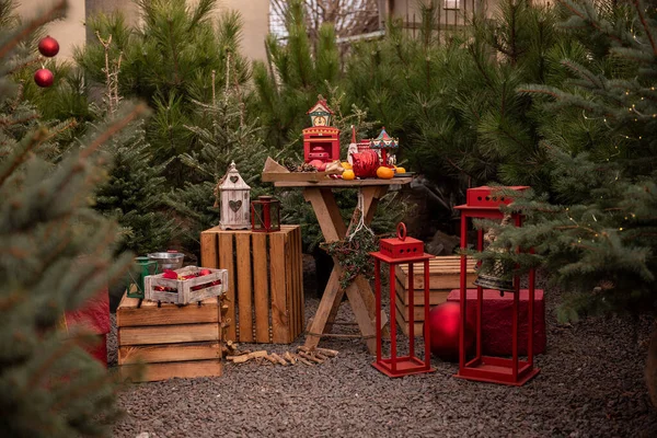 Festive location for decorating outdoor Christmas tree market in city. Holiday fair, backyard decoration. Trendy red color for lanterns, candlesticks, toys. Wreath on wooden table. New Years exterior