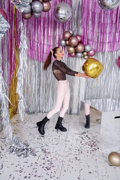 Dancing crazy Fashion girl with beauty makeup, in soft pink pants, mesh bodysuit among confetti, silver shiny foil tinsel. Models having fun with glitter balloons. New Year party. Masquerade party